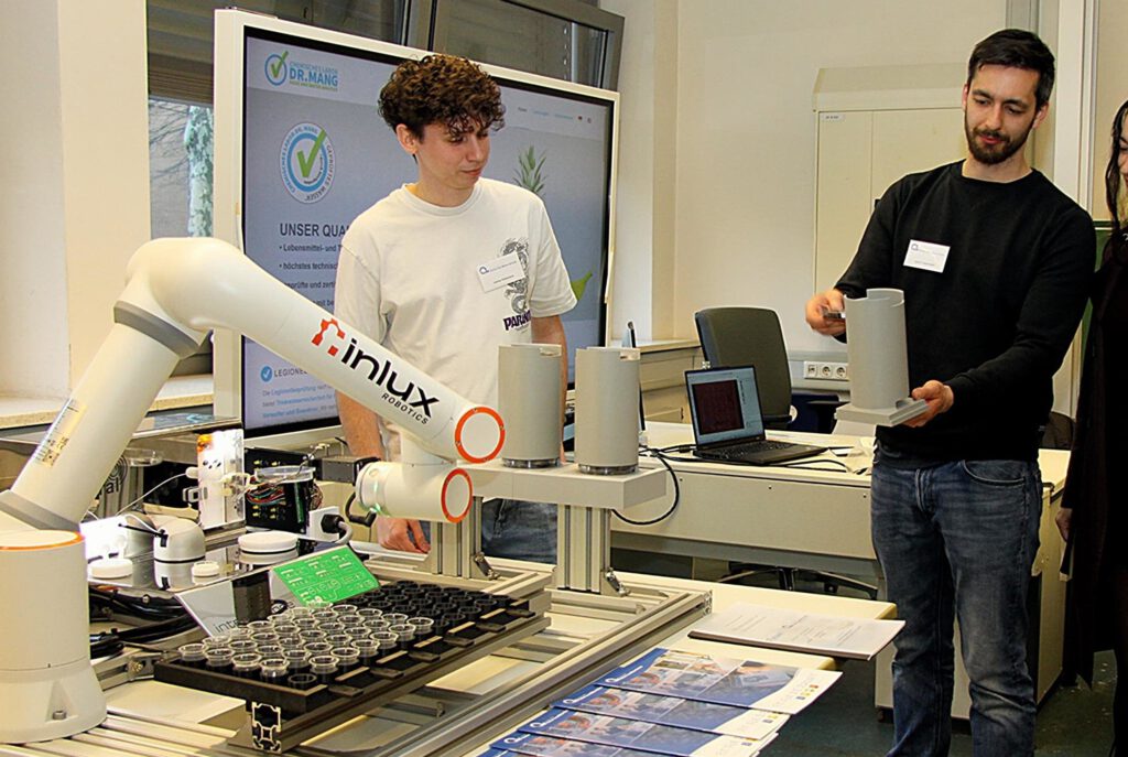 Joshua Kampmann und David Trieschmann demonstrieren einer Messe-Besucherin ihr Projekt der „Automatisierung eines Spiralplaters“