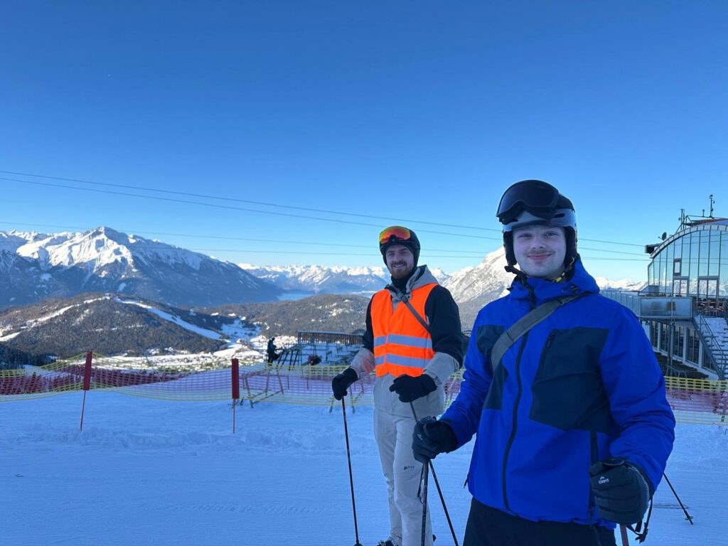 Zwei Teilnehmende in Skiausrüstung stehen bei blauem Himmel an der Bergstation, dahinter die Tiroler Berge.