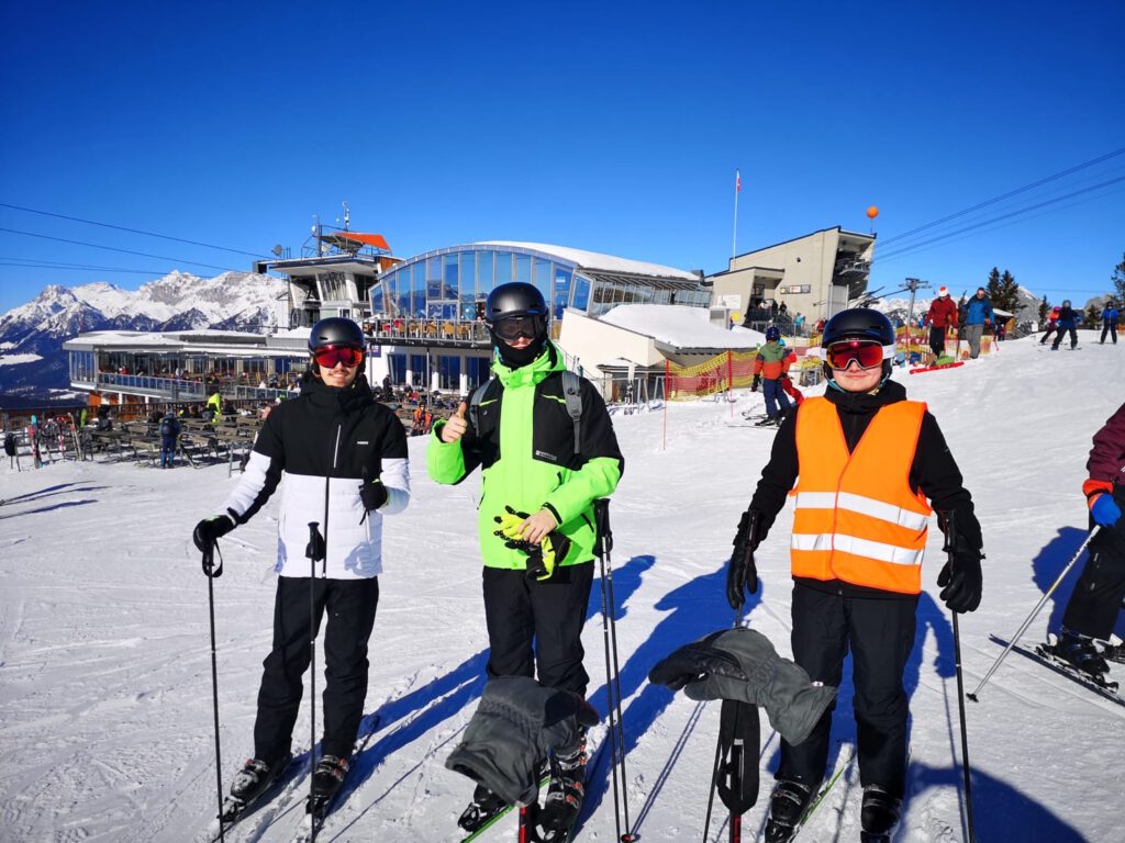 Drei Schüler mit Ski, Helm und Skibrille stehen im Sonnenschein vor der Bergstation.