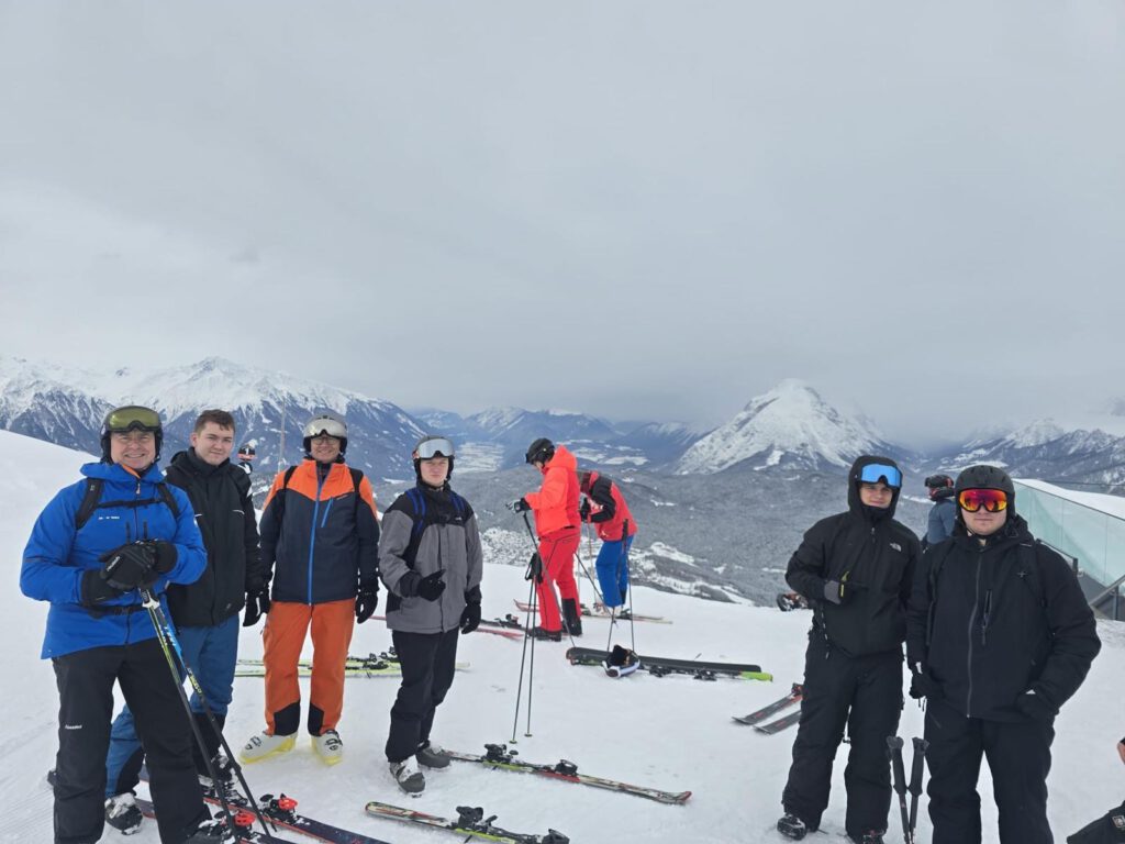 Eine größere Gruppe von Schülerinnen und Schülern steht mit Ski am Pistenrand vor einem Bergpanorama.
