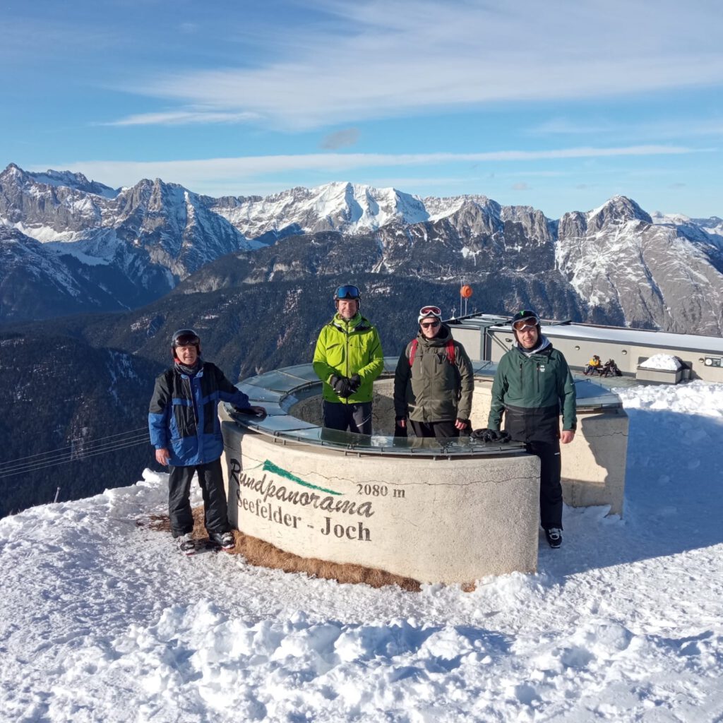 Vier Personen stehen an der Panoramatafel am Seefelder Joch auf 2080 Metern Höhe, dahinter schneebedeckte Gipfel.