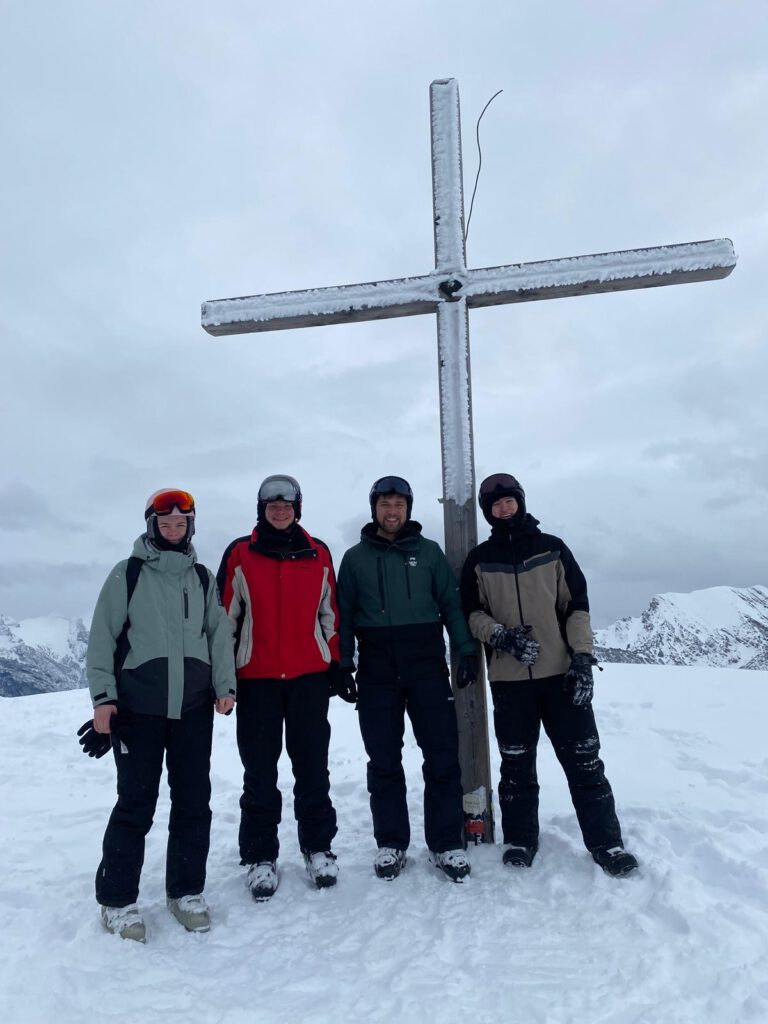 Vier Teilnehmende in Winterkleidung stehen bei bedecktem Himmel vor einem Gipfelkreuz im Schnee.