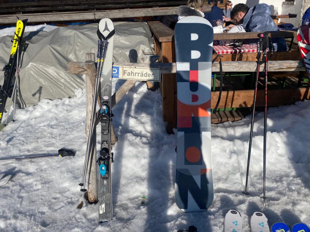 Ski, Skistöcke und ein Snowboard stehen im Schnee vor einer Skihütte.